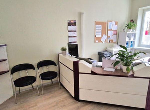 Heller Empfangsbereich einer Arztpraxis mit modernem weißem Empfangstresen, schwarzen Besucherstühlen und grünen Pflanzen am Fenster