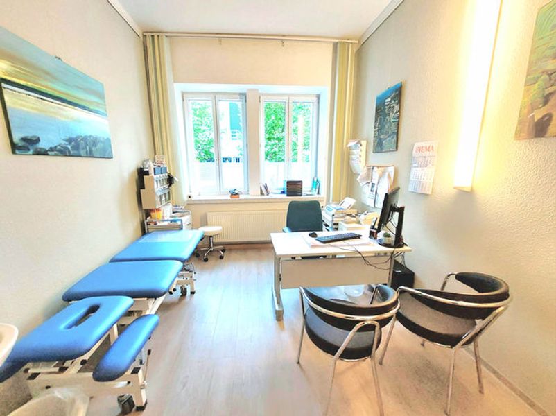Heller Behandlungsraum mit blauer Behandlungsliege, weißem Schreibtisch, Stühlen und großem Fenster mit Blick ins Grüne