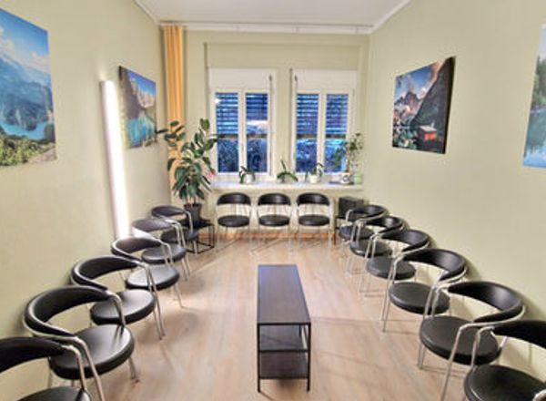 Wartezimmer mit schwarzen Stühlen im Kreis angeordnet, große Naturbilder an den Wänden und Fenster mit blauen Jalousien