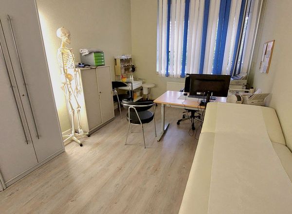 Heller Behandlungsraum mit weißer Untersuchungsliege, Anatomie-Skelett, Schreibtisch mit Computer und blau-weiß gestreiften Vorhängen